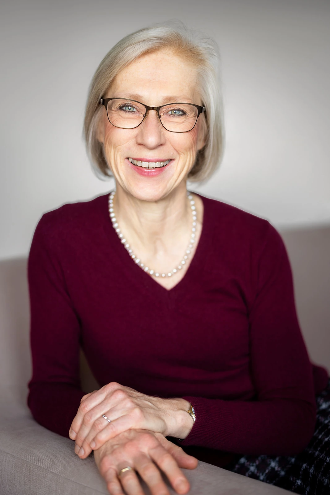 Ältere Frau mit Brille und Perlenkette lächelt freundlich auf einem Sofa sitzend.